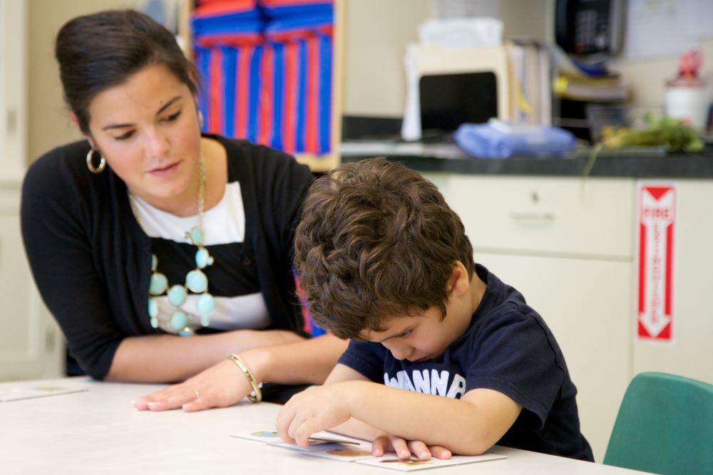 A teacher watching a young boy working on a difficult assignment.