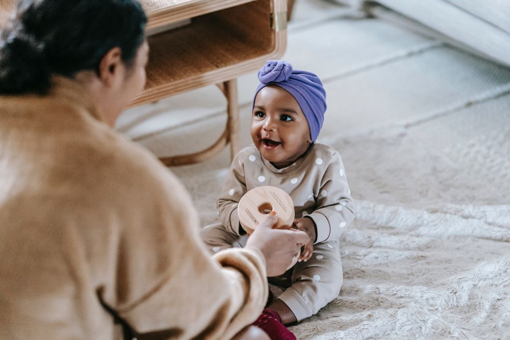 Looking over the shoulder of a woman showing a wooden spool to a toddler.