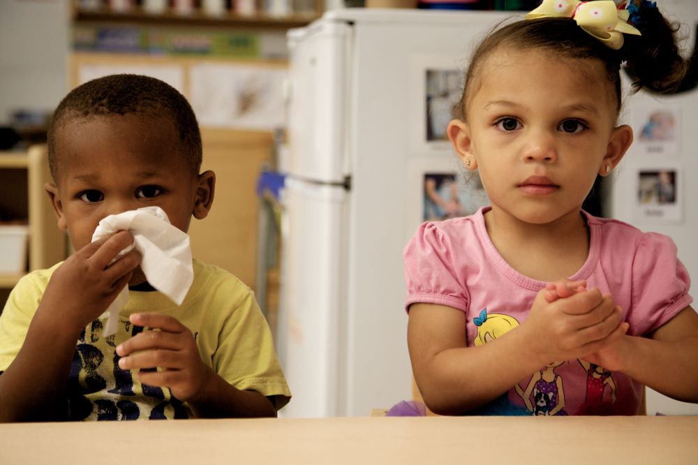 A boy using a tissue to blow his noise, while a girl beside him is washing her hands.