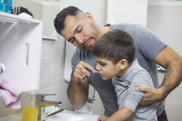 Padre sosteniendo a su hijo para que alcance el lavamanos y pueda cepillarse los dientes.