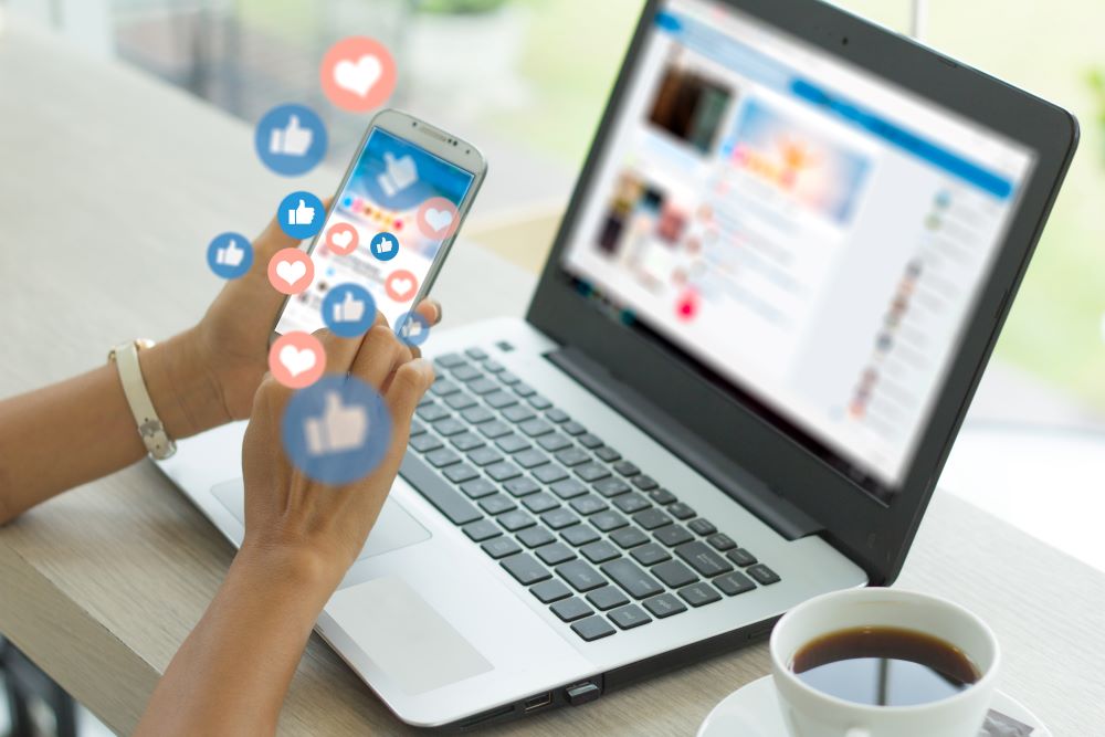 A view of a person holding a smartphone in front of a laptop screen with several floating social media style like and love icons.