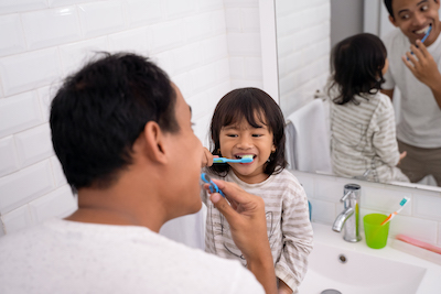 Padre de pie con su hija pequeña sentada en el lavamanos, ambos cepillándose los dientes.