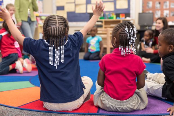 Children gathered on the floor in a circle with one of them holding their arms up high.