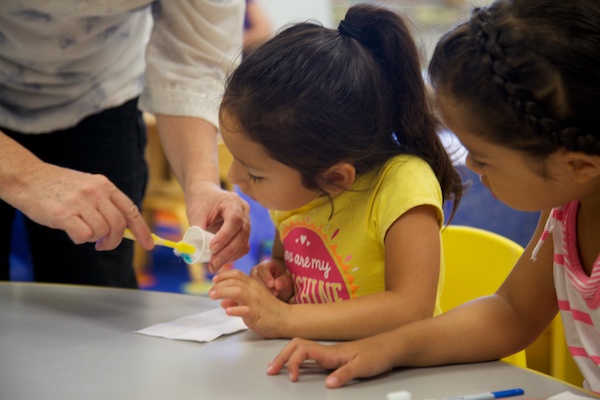 Children being shown how to apply toothpaste to a toothbrush.