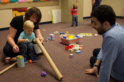 Adults helping infant explore a long cardboard tube.