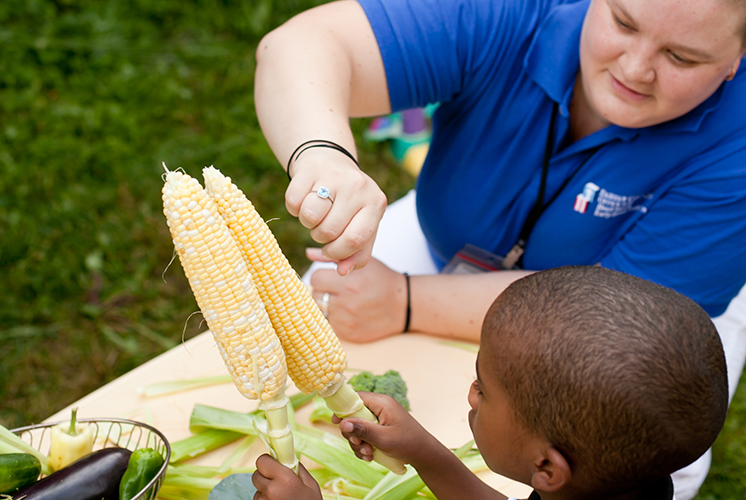 a boy holds ears of corn at a table with a teacher