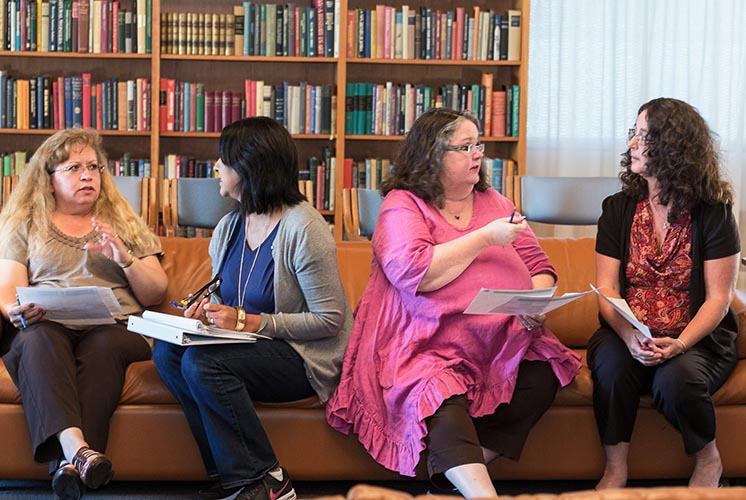 Four women sit on a couch near bookshelves, talking to each other.