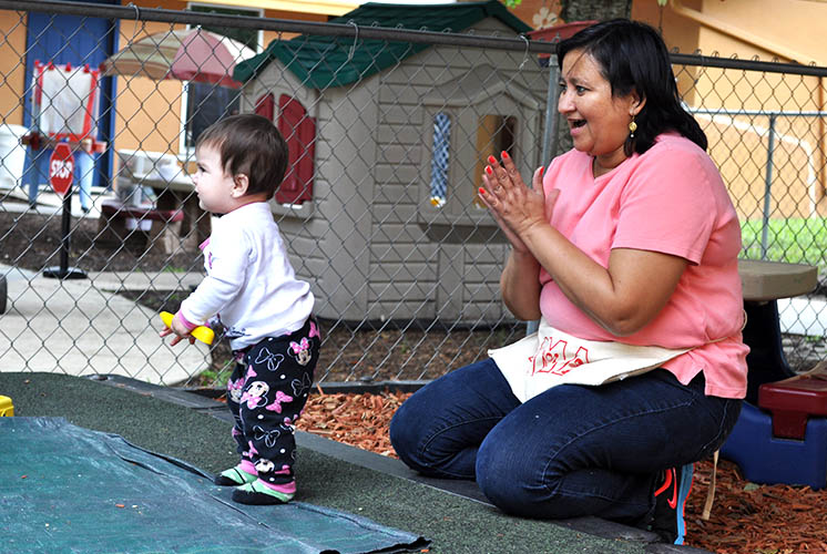 a woman encourages a toddler standing on turf outdoors