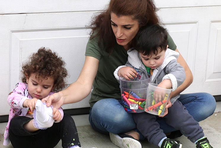 A woman helps a small girl put on socks while holding a playing boy in her lap