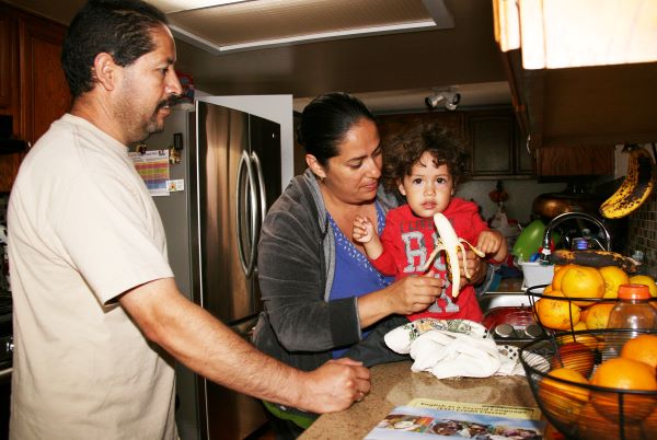 Una madre y un padre en la cocina ayudando a su hijo pequeño a comer un plátano de manera segura.