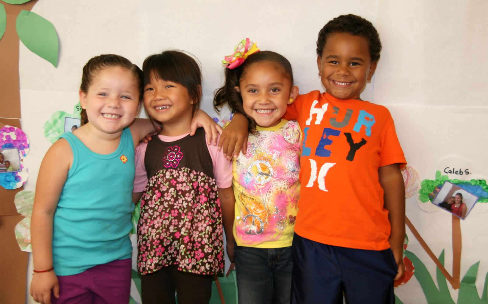 Four young children standing side by side with arms on one anothers shoulders.