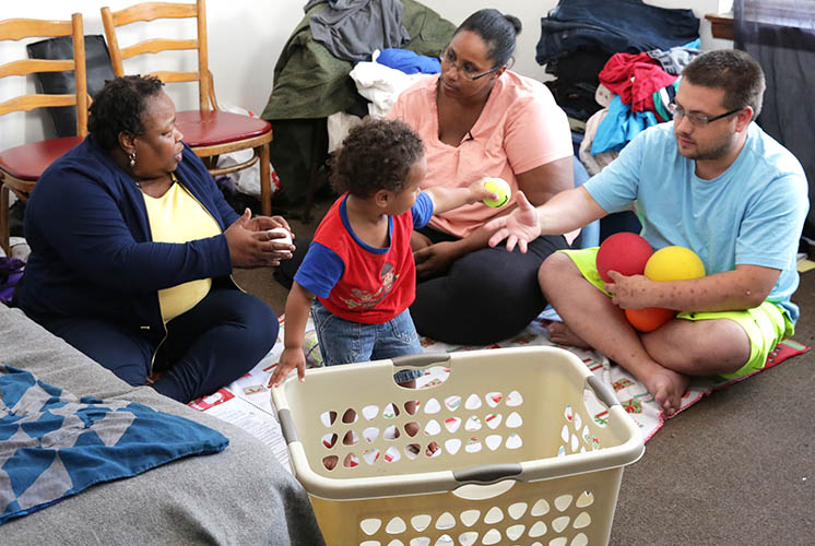 a family sits on the floor around a toddler standing with the help of a laundry basket