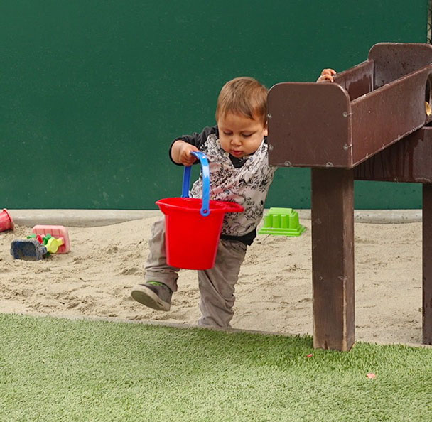 a toddler is carefully stepping out of a sandbox holding a sand pail