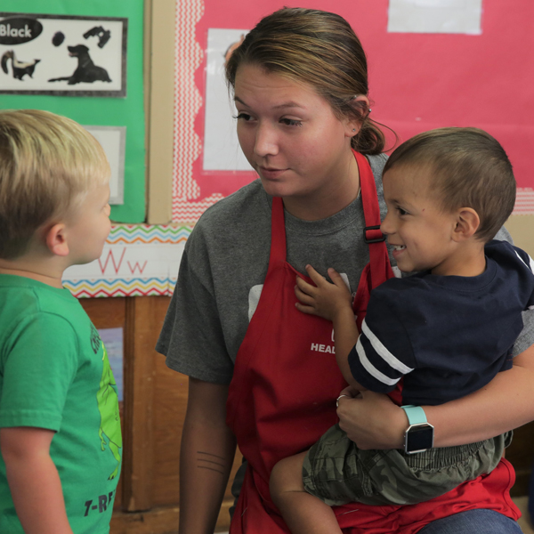 Head Start teacher holding one toddler boy while she talks to another