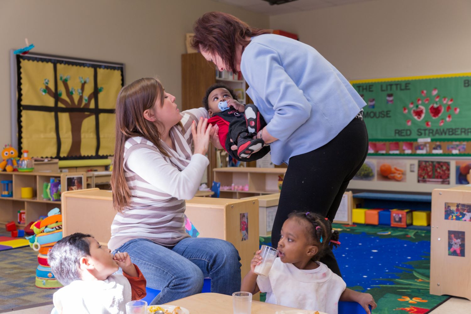 Teacher cradling and handing over a toddler to another teacher.