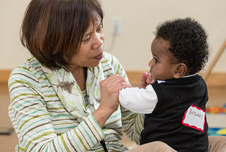 a teacher focuses closely on a toddler, helping him stand